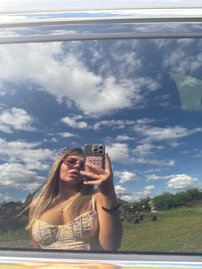 A young woman with blonde hair and pink sunglasses takes a selfie in a car mirror, reflecting a bright blue sky with fluffy white clouds. She is wearing a yellow top with a floral pattern and a gold cross necklace. Her right hand holds a smartphone with a pink pop socket attached to the back. In the background, reflected in the mirror, is a rural landscape with houses, trees, and a field. The image appears to have been taken outdoors on a sunny day.