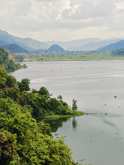 The image displays a serene vista of Rupakot, Nepal, dominated by a vast, calm lake reflecting the hazy sky. The foreground is lush with dense, vibrant green foliage, creating a natural border that spills towards the water's edge. Beyond the immediate greenery, a gently sloping hillside is visible, dotted with a scattering of small houses and buildings, suggesting a rural settlement.
Further in the distance, the landscape transitions into rolling hills and majestic mountains, their peaks softened by atmospheric haze. These layers of mountains recede into the horizon, painted in muted shades of blue and green, under a sky filled with soft, diffused clouds. The lake itself is dotted with a few small, dark objects, possibly aquatic plants or debris, and a larger, dark shadow stretches across its surface, hinting at the presence of a boat or a significant object below the water. A single red structure, likely a building or boat dock, is situated on the edge of the lake, partially obscured by the trees. The overall impression is one of tranquility and natural beauty.