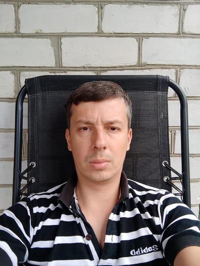 A medium shot shows a man sitting in a black outdoor chair with a white brick wall behind him. The man is looking directly at the camera with a neutral expression. He has short, dark hair with some graying at the temples, and brown eyes. He is wearing a black and white horizontally striped polo shirt with a dark collar. The Adidas logo is visible on the lower right side of his shirt. A thin gold chain is visible around his neck. The lighting suggests it is daytime, and the setting appears to be outdoors, possibly a patio or backyard given the chair and brick wall. The image is an informal selfie.