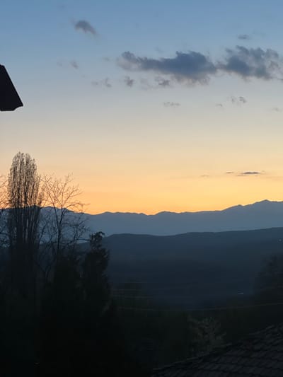 The image captures a landscape at sunset or sunrise, likely in Kribul, Bulgaria. The sky transitions from a pale blue with scattered gray clouds to a golden-orange hue. Silhouetted mountain ridges are visible against the lighter sky. In the foreground, bare tree branches form a dark, intricate pattern. Power lines are also visible. The very bottom of the frame has a roof. The image is rotated.