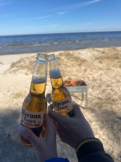 The image shows two hands holding bottles of Corona Extra beer in front of a beach scene. In the background, there is a fire pit with a small fire burning. The beach sand is visible, as is a body of water under a blue sky. The location is possibly Zīvartiņš, Latvia, based on the OCR text on the beer bottles. One hand is wearing a jacket with a black and yellow striped cuff, while the other hand shows fingernails painted white with a lilac base. The bottles are clinking in a cheers.