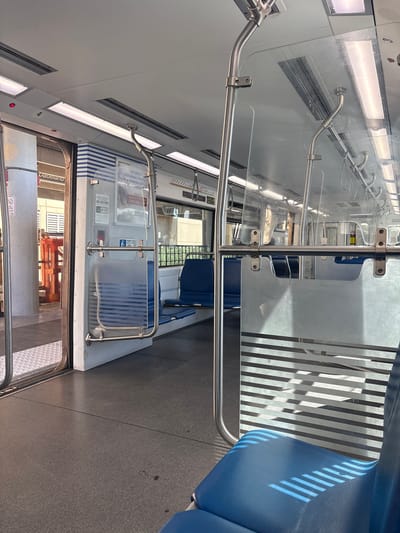 The image shows the interior of a Metrorail train car in Kendall, Florida. The train car is empty. Blue seats are visible, some with sunlight streaming through a patterned partition, casting striped shadows. There are handrails and overhead lighting. To the left, the doors are open, revealing a station platform with a pillar and orange scaffolding. A sign on the pillar reads "Dadeland".