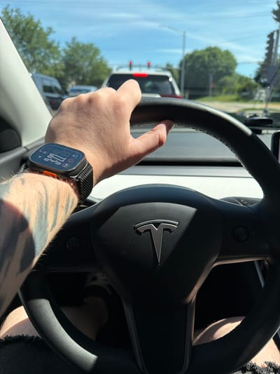 A first-person perspective shot from inside a Tesla car shows a hand gripping the steering wheel. The hand is on the left side of the frame and features a smartwatch on the wrist. The car is in traffic, with other vehicles and traffic lights visible through the windshield. Trees and a blue sky with clouds are in the background. The scene is likely in Buffalo Grove, United States of America, given the visual cues typical of suburban areas in that region.