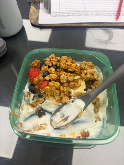 A top-down, close-up shot shows a partially eaten container of yogurt with granola, fruit, and a dark gray spoon resting inside. The yogurt is white, and mixed with the granola are blueberries, raspberries, and banana slices. Some of the granola and fruit are coated with a brown substance, likely peanut butter or a similar nut butter.

The container is square with rounded corners and appears to be transparent with a light green tint. The container rests on a dark gray surface that has hints of silver. To the left of the container is a clipboard holding a form with printed text in French. A red pen rests on top of the clipboard. The lower left of the frame shows a gray water bottle and the top of a gray cap.

The scene appears to be outdoors, as indicated by natural lighting and shadows. The location, Longueuil, Canada, is based on available information. No people, subjects, interactions, or environment details beyond the immediate setting are visible.