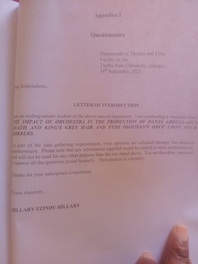 This media file is a close-up, overhead shot of a document, likely a questionnaire or letter of introduction, lying on a surface. The document is printed in black text on white paper, with some sections highlighted or in larger font. The image's focus is on the content of the document, with a portion of a person's hand, including a thumb and index finger, visible in the lower right corner, suggesting someone is holding or pointing to the paper.

The text on the document includes "Appendixes 2," "Questionnaire," and "LETTER OF INTRODUCTION." It details a research being conducted by an undergraduate student from the Department of Theatre and Film, Faculty of Art, Taraba State University, Jalingo. The research topic appears to be related to "THE IMPACT OF ORCHESTRA IN THE PRODUCTION OF DANJA ABDULLASS DEATH AND KING'S GREY HAIR AND FEMI OSOFISAN'S ONCE UPON FOUR ROBBERS." The letter explains that opinions are solicited through a questionnaire and that information supplied will be treated with strict confidentiality. It also states that participation is voluntary. The document is dated "14th September, 2025" and is signed "HILLARY EZINDU HILLARY."

The scene is indoors, with soft lighting that casts a slight pinkish hue over the paper, suggesting ambient lighting rather than direct sunlight. The overall impression is of a formal or academic context, given the nature of the document. The presence of the hand indicates a human interaction with the document, possibly during the process of reading, filling out, or presenting it. The location context provided (Magajin Gari, Nigeria) helps to place the origin of this document within a specific geographical and cultural setting, likely related to the university mentioned.