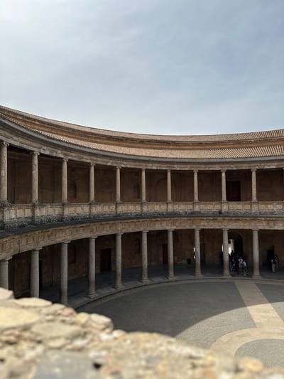 The image displays a grand architectural scene, capturing a circular or semi-circular courtyard of a historic building, strongly resembling the Palace of Charles V in Granada, Spain. The perspective is slightly elevated, looking across and down into the expansive courtyard.
The main subject is the impressive multi-story arcaded structure that defines the perimeter of the courtyard. It features two visible tiers of arches supported by numerous sturdy stone columns. The stonework exhibits intricate details and patterns. To the right, the curved exterior wall or roofline of the building is visible against the sky, showing a textured surface.
On the ground level of the arcade, several small groups of people are scattered, likely tourists exploring the site. They are seen standing and observing, adding a sense of scale to the massive architecture. The courtyard floor in the foreground is paved with large, textured stones arranged in a decorative pattern, displaying distinct areas of light and shadow, suggesting bright sunlight. A blurry, light-colored stone wall or balustrade occupies the very bottom right foreground, indicating the viewpoint from a barrier or opening.
The sky, visible in the upper left, is clear and bright blue, suggesting good weather conditions. The strong shadows cast by the columns and arcades across the paved ground indicate that it is daytime, possibly midday or early afternoon. No visible text is present in the image.
