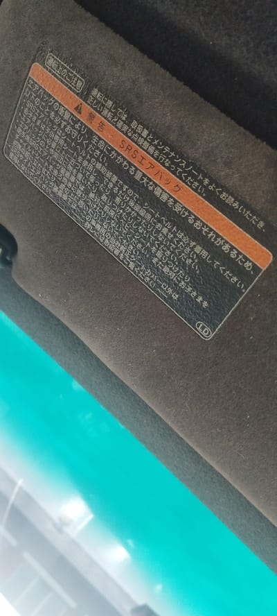 The image is a close-up, high-angle shot of a car's sun visor, which is covered in a dark, fuzzy fabric. A sticker is affixed to the visor. The sticker has an orange header with Japanese text that translates to "Caution." Below this, in larger Japanese text, are warnings and instructions regarding the SRS airbag system, with the words "SRS Airbag" highlighted in red. The sticker also contains a circular emblem with the letters "LD" inside. The background of the image is out of focus and shows a bright, turquoise surface, likely the sky or a reflection. The location in the city of Покосное, Russia is not visible or inferable from the image.