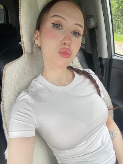 A young Caucasian woman with long brown hair braided down her right side is sitting in the passenger seat of a car. She is wearing a white t-shirt and has heavy eye makeup with winged eyeliner and bright pink blush. Her lips are pursed, and she is looking directly at the camera. The car interior is visible, including a tan fuzzy seat cover, a black seatbelt, and a window showing a green, tree-lined outdoor environment. The location is identified as Gavrilovka, Russia.