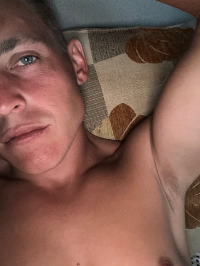 This is a close-up, overhead, indoor shot of a fair-skinned individual with short brown hair, lying down and looking towards the camera. The individual is shirtless, revealing their chest and armpit. They have blue eyes and are lying on a patterned surface that appears to be a bed or couch, with a blue and brown fabric. The lighting suggests it is daytime. There is no information in the image to indicate the location is Żary, Poland.
