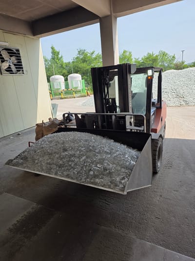 A Doosan forklift is parked indoors under a concrete overhang. The forklift's hydraulic lift is lowered, holding a large metal bucket filled with clear glass shards. The glass shards appear to be small, cut pieces of bottles. In the background, there are two large white storage tanks with green fencing around them. Beyond the tanks, there are trees and a large pile of similar glass shards in an outdoor area. The ground is concrete, and a large tan bag is visible to the left of the forklift. The scene is likely at a glass recycling facility in Anseong-si, South Korea.