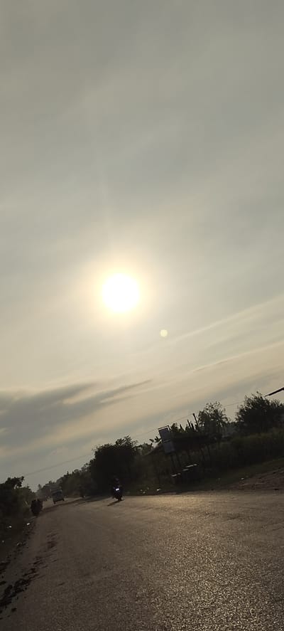 The image shows a road with vehicles and trees under a bright, hazy sky. A sun is visible in the upper portion of the image, with a smaller orb below it, possibly a reflection or another celestial body. The road appears to be asphalt and has some wet patches, reflecting the light. On the side of the road, there are trees and vegetation. Several vehicles are present, including a motorcycle in motion, a truck, and possibly other cars and people further down the road. There is a simple structure, possibly a bus stop or shelter, with some signage on the right side of the image. The overall impression is a rural or semi-rural road scene in Sumbokrayek, Indonesia, likely during the daytime.