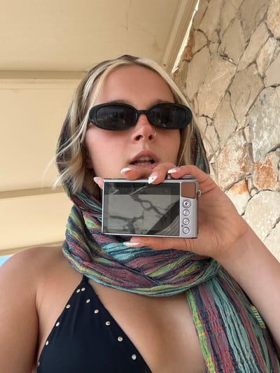 A woman is taking a selfie with a digital camera. She is wearing a colorful scarf wrapped around her head and neck, black sunglasses, and a black bikini top. The background shows a stone wall and a cream-colored structure, suggesting an outdoor setting. The lighting indicates it is daytime. The woman appears to be smiling or laughing. The camera she is holding is silver with brown accents and several buttons on the back. The image is oriented vertically, as if the woman is holding the camera up to take the photo herself.