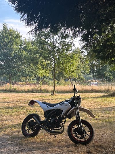 A white dirt bike with red trim on its wheels is parked on a dry grassy field. The bike is facing slightly to the left. In the background, there are trees with green leaves, and a sign that reads "SORTIE PUBLIQUE". The sky is blue with some clouds. The scene appears to be in an outdoor, rural setting.