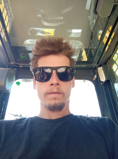 The image is a selfie taken from the perspective of someone inside the cab of a construction vehicle. The subject, a young man with light brown, tousled hair and a goatee, is wearing sunglasses and a black t-shirt. The interior of the cab is visible, with a perforated headliner and what appears to be a warning label with the number "85" printed on it. The exterior environment is bright and appears to be daytime, with hints of greenery visible through the windows. The location of Preiļi, Latvia, is not evident from the visual information in the image.
