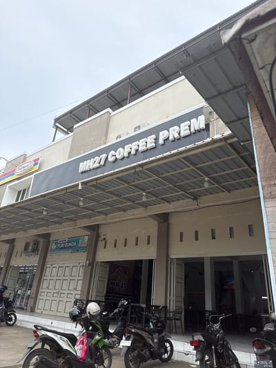 The image shows the facade of a commercial building in Meunasahmanyet, Indonesia, on an overcast day. The building features a prominent sign that reads "MH27 COFFEE PREM" in white lettering against a dark gray background. To the left of the coffee shop sign, there is an "Indomaret" convenience store sign. Below the main sign, a covered walkway extends outwards, supported by metal beams and a corrugated metal roof. This walkway is illuminated by several light bulbs.

In front of the building, there is a row of parked motorcycles. Some people are visible, including a person on a motorcycle in the lower left corner and other individuals in the background inside the coffee shop. The coffee shop entrance appears to be open, revealing tables and chairs inside. To the left of the coffee shop entrance, there is a separate storefront with a sign that reads "APOTEK PURI BUNDA". The building's exterior is painted in neutral tones, with rectangular window-like openings visible on the upper floors. The ground in front of the building is paved.