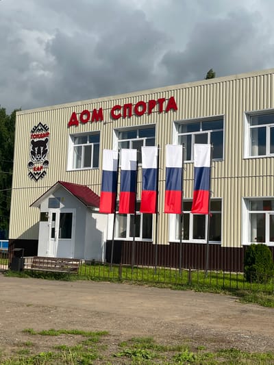 The image shows a building with the words "ДОМ СПОРТА" in large red letters. The building is beige with vertical corrugated siding. To the left of the entrance, there is a crest with a bear and some text. Three vertical Russian flags are displayed in front of the building. There is a small, white structure with a red roof to the left of the entrance, likely a ticket booth or small office. The area in front of the building is a dirt or gravel path with a small metal fence and some grass. In the background, trees are visible. The sky is overcast. The scene appears to be a sports facility.