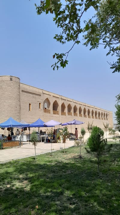 The image depicts an outdoor marketplace in Denov, Uzbekistan, in front of a long, tan brick building with arched openings. Several vendors are set up under blue and purple tents, displaying goods that appear to include books and clothing. Some people are browsing or interacting with vendors, while others are walking through the market area. The ground is paved in some sections and covered with grass in others, with several small trees and bushes scattered around. Overhead, the branches of a large tree with green leaves partially obscure the clear blue sky. The building itself features a rounded tower on the left side and numerous arched alcoves running along its length, suggesting historical architecture. The overall scene is one of a bustling daytime market in a location with both traditional and natural elements.