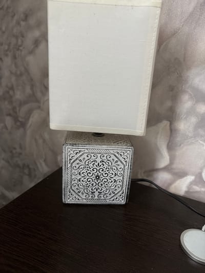 A close-up, eye-level shot shows a small table lamp on a dark wood surface. The lamp has a white fabric lampshade and a square base with an intricate black and white patterned design, resembling traditional tilework. A black power cord extends from the base and disappears to the right. In the background, a textured wallpaper with a floral motif is visible. The overall impression is one of a domestic setting, possibly a bedroom or living room, in Kinel'-Cherkasy, Russia.