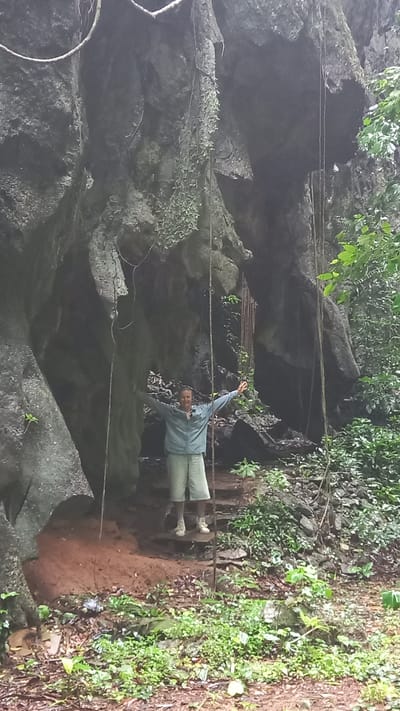 A man in a light blue long-sleeved shirt and light-colored pants stands with arms outstretched in the entrance to a cave. The cave entrance is surrounded by vegetation and rock formations. Ropes are strung across the entrance. The location is Ban Pak Ra, Thailand.