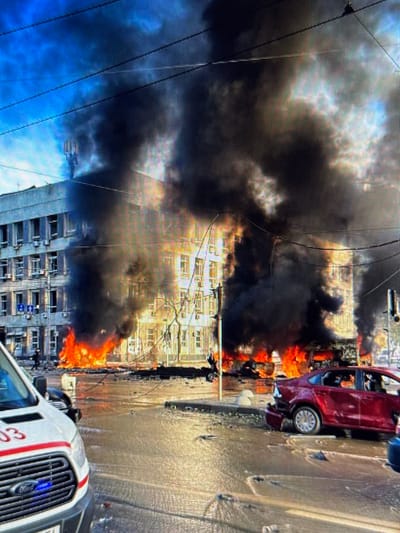 This image captures a scene of destruction in Shcherbanka, Ukraine, with a large building engulfed in flames and thick black smoke billowing into the sky. The fire appears to be concentrated on the lower floors of the multi-story building, with bright orange flames visible through the windows and emerging from street level. Debris litters the ground, and the street is partially covered in water, suggesting a possible aftermath of an explosion or firefighting efforts.
In the foreground, the front of a white emergency vehicle, possibly an ambulance, is visible, displaying the number "03" in red lettering on its side. To the right, a damaged red sedan is parked on the street, its rear end showing signs of impact. The overall environment suggests a chaotic and dangerous situation, likely caused by an attack or accident. Power lines are visible overhead, adding to the urban setting. The sky is a mix of blue and dark clouds, partially obscured by the smoke.