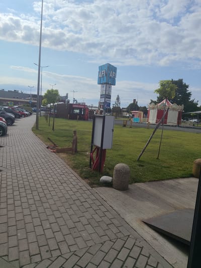 This image depicts an outdoor scene with a sidewalk in the foreground, leading to a grassy area with a few trees. In the background, there are several buildings, including what appears to be a shopping center or commercial complex, and a few cars are parked along the sidewalk. There is a tall sign with the letters "AFI" prominently displayed, which likely signifies the AFI Palace Arad shopping mall. Further into the scene, there are some portable food stalls or kiosks, one with the text "CÂRNAT VINEZ" visible, suggesting a food vendor. A small, festive tent, possibly for an amusement or event, is also present in the middle ground. The sky is partly cloudy. The overall environment suggests a commercial or recreational area in Arad, Romania, likely during daylight hours. There are no people clearly visible in the image.