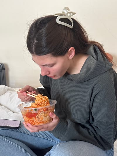 An indoor scene, set in what is identified as Huangdao District, China, captures a young female engaged in eating. The female, appearing to be in her late teens or early twenties, has dark brown hair pulled back with a light-colored, abstractly shaped claw clip. She is dressed in a dark grey, ribbed long-sleeve hooded top and blue denim jeans. She is seated, likely on a bed covered with white bedding, and is actively lifting noodles from a clear plastic bowl with light-colored chopsticks. The noodles are coated in a reddish-orange sauce and contain visible pieces of tomato and other ingredients. To her immediate left, a smartphone with a dark case rests on the white surface, displaying content on its screen. In the background, a portion of a dark-colored, hard-shell suitcase is visible against a plain, light-colored wall.