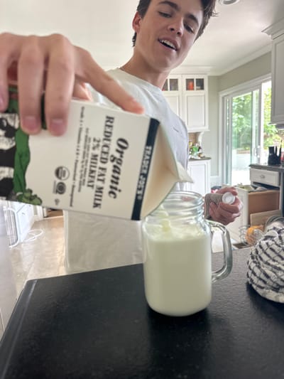 A young person holds a carton of Trader Joe's Organic Reduced Fat 2% Milk. The carton is in focus, and its details are legible. Behind the carton, another person pours milk from a second carton into a clear glass mug. The mug is nearly full. The scene is inside a kitchen with white cabinets, black countertops, and a window showing greenery outside. A patterned cloth is partially visible near the mug.