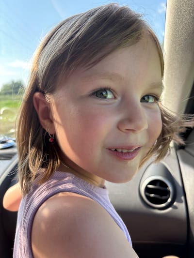 The image is a close-up portrait of a young girl with brown hair and green eyes, seen from the chest up, in what appears to be the interior of a car. She is wearing a purple tank top and a small dangling earring in her left ear. Sunlight illuminates her face from the side, casting a warm glow. The background is blurred, but hints of greenery and a bright sky suggest an outdoor environment. A car's air vent is visible on the right side of the frame. The girl is looking towards the right of the camera, with a slight smile. The scene is devoid of any context that would place it in the city of Маркидоновка, Russia; no landmarks or identifying features of that specific location are present.