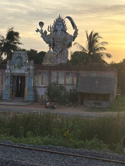 This image depicts an outdoor scene at dusk in Pachayankuppam, India. The primary subject is a large, multi-armed statue of a deity, likely Hindu, with intricate ornamentation. The statue is positioned in front of a weathered building with a thatched roof. To the right of the statue and building, tall grass and lush green vegetation are visible, bordering a railway track made of gravel and metal rails. The sky is filled with soft, diffused light, indicating either sunrise or sunset, with a warm, golden hue.

The building appears to be a temple or shrine, with architectural details and a smaller deity statue within an alcove above the entrance. A motorcycle is parked near the entrance of the building, suggesting recent activity or local transportation. The overall atmosphere is serene, with the fading light casting a tranquil mood over the scene.

Visible text is present on a sign affixed to the building, written in Tamil script. While the exact translation is not provided, it likely contains information related to the temple, its deity, or visiting hours. The scene is set in a rural or semi-rural area, characterized by the natural vegetation and the presence of the railway line.