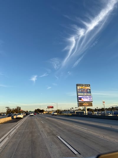 A sunny highway scene with a clear blue sky dotted with wispy, feather-like cirrus clouds. Multiple billboards are visible, advertising Chargers tickets and GED programs. Cars are present on the right-hand side of the image. The photograph is taken from the perspective of someone traveling on the highway.