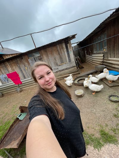 A woman with long brown hair and a black t-shirt with white polka dots takes a selfie in an outdoor courtyard in the village of Baldeyka, Russia. She is smiling at the camera. In the background, several white ducks are gathered around shallow bowls and a blue plastic basin. Behind the ducks are rustic wooden structures, possibly sheds or animal enclosures, with corrugated metal roofing. Laundry, including a red shirt and a grey garment, hangs on a clothesline. The ground is dirt and mud, with patches of green grass. The sky is overcast and grey.