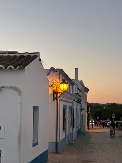 Captured from a ground-level perspective, the image showcases a street in Vila Real de Santo António, Portugal, during twilight. A row of white buildings with blue accents extends into the depth of the image, running parallel to a pedestrian walkway. A bright orange light fixture affixed to one of the buildings illuminates the immediate surroundings. A group of pedestrians and parked vehicles are visible further down the street. The sky transitions from a gradient of orange near the horizon to a pale grey closer to the top of the frame. The buildings are predominantly white with tiled roofs.