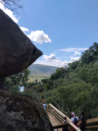 The image shows a landscape view of Alijó, Portugal, featuring a large granite boulder dominating the left side of the frame. To the right of the boulder, a wooden walkway with handrails leads downhill. Several people are on the walkway, including a group of three in the foreground and another person slightly further back. The vegetation includes trees and shrubs on the right and in the background. In the distance, rolling hills and a river can be seen under a blue sky with scattered clouds. Power lines stretch across the sky in the upper right portion of the frame.