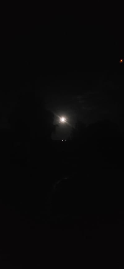 The image captures a nighttime scene with a bright moon illuminated against a dark, cloudy sky. In the foreground, silhouetted against the moonlight, are dark shapes that appear to be trees and possibly buildings. Two small, bright lights are visible in the lower middle of the frame, suggesting artificial illumination, perhaps from streetlights or vehicles. To the far right, a small, reddish-orange object is present in the sky. The overall impression is a dimly lit, possibly rural or suburban environment at night, with the moon being the dominant light source. The location in Veshenskaya, Russia is not explicitly depicted in the visual elements of the image, as there are no identifying landmarks or contextual clues.
