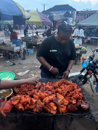 In Obinze, Nigeria, a woman stands in front of a large grill filled with raw, marinated meat. She wears a black t-shirt with "YN STUDIO" printed on it and several bracelets. Her hands are positioned near the meat, as if preparing to cook it. The scene takes place outdoors, with several market stalls and umbrellas visible in the background. A motorcycle is parked to the right of the woman, with its headlight illuminated. The ground appears to be dirt and debris, typical of an outdoor market. Other people are present in the background, some obscured by umbrellas. The sky is overcast, suggesting it might be early morning or late afternoon.