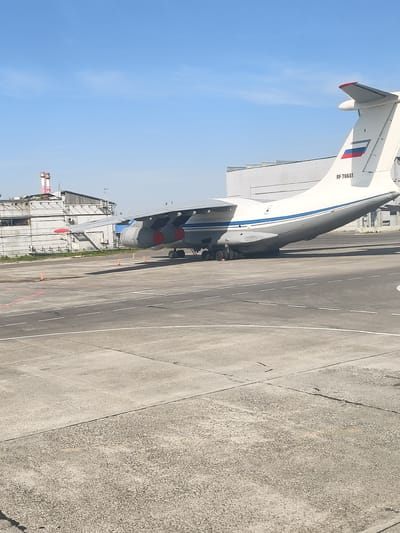 An Ilyushin Il-76 aircraft, registration RF-78681, is parked on the tarmac at Emelyanovo Airport (УККК/УТКК) in Krasnoyarsk Krai, Russia. The aircraft is white with a blue stripe along its fuselage and a Russian flag on its tail. It has four turbofan engines mounted on the wings. In the background, there is a light-colored industrial building with two tall chimneys. The sky is clear and blue. The tarmac is concrete with markings. There are several orange traffic cones visible.