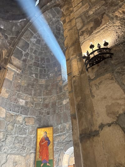 A stone arched ceiling is illuminated by a shaft of sunlight. An ornate black metal chandelier hangs from the ceiling. A religious icon depicting a figure in red robes is mounted on the wall. The ceiling and walls are constructed of irregularly shaped stones, creating a textured surface.