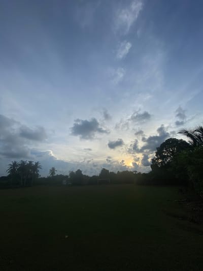 This image shows a wide, dark green, open field, likely a sports field, with a soccer goal visible on the left side and another on the right in the distance. The field is bordered by a dense line of trees and vegetation, with several palm trees scattered among them on the left. A small, white building is partially visible behind the trees in the center. The sky above is filled with dramatic, dark gray clouds interspersed with patches of bright light, suggesting either sunrise or sunset. The sun itself is a small, bright orb peeking through a gap in the clouds on the right side of the frame, casting a warm glow on the cloud formations. The overall impression is of a somewhat overcast and atmospheric evening or morning in a tropical or subtropical location. The specific location of Keudeublang, Indonesia, is not visually identifiable from the image content alone.