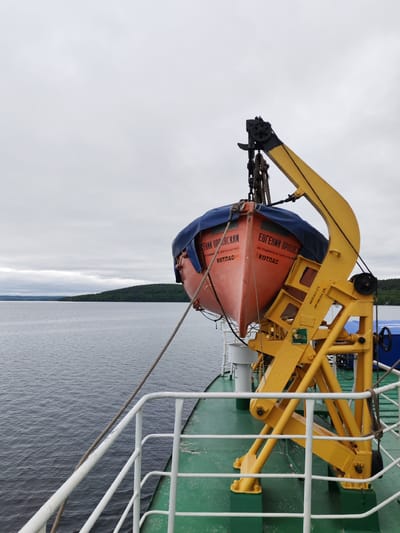 An outdoor wide shot from the deck of a vessel captures a tranquil waterway scene under an overcast sky. In the immediate foreground, a bright orange lifeboat, partially covered with a blue tarp, hangs suspended from a sturdy, yellow metal davit. The lifeboat displays Cyrillic text: "ЕВГЕНИЙ ОРЛОВСКИЙ" (Yevgeny Orlovsky), "АО РЫБИНСКОЕ ПАРОХОДСТВО" (Rybinsk Shipping Company), and "КОТЛАС" (Kotlas), indicating its name, operator, and home port. The yellow davit also has markings "ИСП.04.24" (Inspection/Expiry April 2024) and "ПРОВ.04.26" (Test/Next Test April 2026), suggesting maintenance records.

The green deck of the vessel, with white railings, is visible in the lower portion of the image. The vessel navigates a wide body of calm, dark water that extends to a distant shoreline blanketed by dense green forest. The sky is uniformly cloudy and grey, creating a soft, diffused light over the scene. No people are visible. The setting is likely a large lake or wide river, consistent with the provided location context of Medvezhyegorsk, Russia. The lifeboat's position suggests it is either ready for immediate deployment or recently returned from use/maintenance.