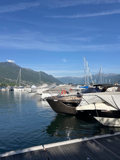 The image shows a marina in Locarno, Switzerland, with several boats docked along a wooden pier. The boats are mostly covered, suggesting they are not currently in use. Mountains rise in the background, and the sky is clear with a few contrails. The water is calm, reflecting the boats and surrounding scenery. The scene is bathed in sunlight, indicating it is daytime. The focus is on the docked boats, with the surrounding environment providing context.