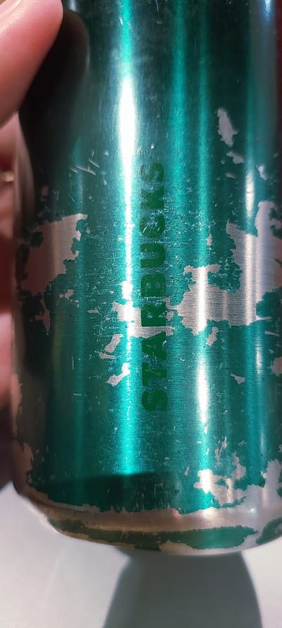 The image shows a close-up of a green metallic can, likely a thermos or a beverage container. The can has the word "STARBUCKS" printed vertically in green lettering, but the outer coating is chipped and peeling, revealing the silver metal beneath in several areas. The lighting creates strong vertical reflections on the can's surface. A person's fingertip is visible in the upper left corner, touching the can. The background is out of focus and appears to be a light-colored, possibly white or grey surface. There is no indication of a specific location in Pagedangan, Indonesia, or any other city within the image.