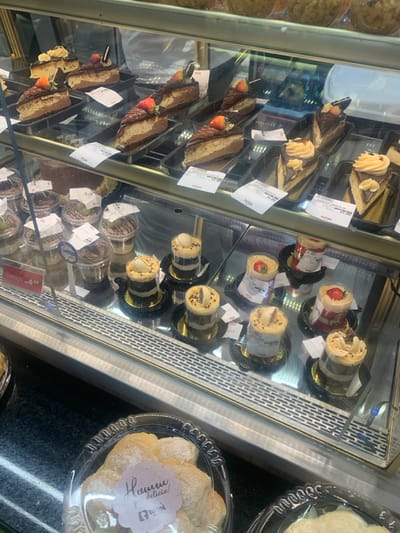 This image captures a refrigerated display case inside Twist Panificadora e Confeitaria, located at Av. Santos Dumont, 1851 - Centro, Lauro de Freitas, Bahia, Brazil.
The scene focuses on a multi-tiered glass display case showcasing various desserts. The top shelf holds multiple slices of cake, primarily chocolate-based with light-colored cream layers, topped with chocolate ganache or icing and a fresh strawberry. Some slices also feature decorative chocolate pieces or a swirl pattern. Several price tags, primarily white with red text, are affixed to the glass in front of these cakes, indicating prices such as R$ 14.90.
The bottom shelf contains numerous individual desserts in clear plastic cups. On the left, a collection of desserts appears to be mousses or layered puddings, some topped with chocolate shavings or green fruit, each with a price tag. One red tag prominently displays "Promoção R$ 4.99." On the right, cylindrical layered desserts, possibly trifles or small cakes, are presented in clear cups on black or gold bases. These vary in appearance, with some featuring white cream or chocolate toppings with a decorative stick, and others appearing to be strawberry-flavored with a red hue.
In the immediate foreground, outside the display case, a clear plastic container holds multiple light-colored, powdered pastries. A pink label on this container reads "Hummm que delicia!" with a small "twist" logo underneath. A portion of another similar container is visible to the right. The display case itself features metal grating along its bottom front edge. Reflections in the glass suggest the presence of a person or a typical store interior. No direct human subjects are visible.