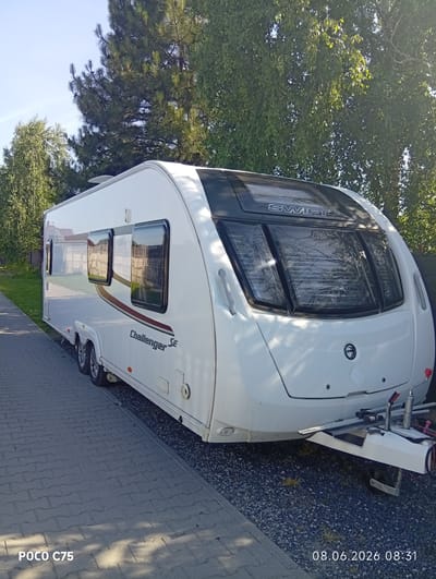 A white Swift Challenger SE caravan is parked on a paved surface in an outdoor setting. The caravan is oriented facing right, with its front end towards the viewer. It is positioned on a pathway made of interlocking grey rectangular pavers. To the right of the pathway, there is an area covered with dark grey gravel. Lush green trees and foliage form a backdrop behind the caravan. The sky is a clear blue. The image captures the caravan from a slight side angle, showcasing its windows and the "Swift" and "Challenger SE" branding. The timestamp "08.06.2026 08:31" and the text "POCO C75" are visible in the lower portion of the image. This scene is located in Kiełczów, Poland.