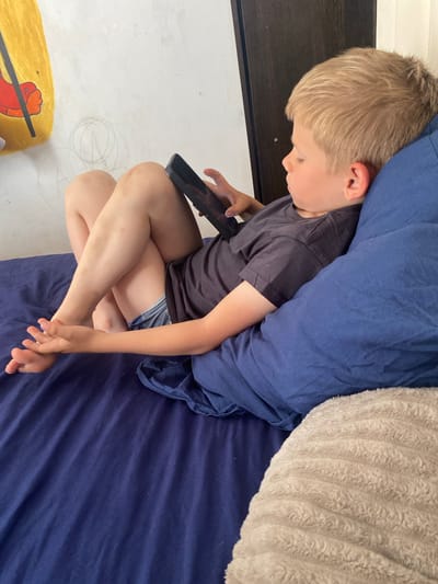 A fair-skinned child with blonde hair is seated cross-legged on a navy blue bed. The child is wearing a dark grey t-shirt and denim shorts. The child is holding a black smartphone. A blue duvet and a light grey pillow are positioned behind the child. The floor is white and marked with pencil drawings and a yellow object with a red design. Based on the information provided, the location is Vasyurinskaya, Russia.