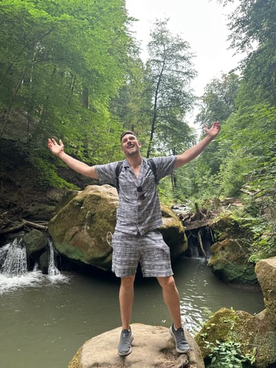 A man stands with his arms outstretched in a lush green forest next to a small waterfall and a body of water. He is wearing a grey patterned short-sleeved shirt and matching shorts, and grey sneakers. He has a backpack on his back and sunglasses hanging from his shirt. The man is smiling and looking up at the sky. The forest is dense with trees and foliage, and the water is clear and reflects the surrounding greenery. The scene appears to be a natural setting, possibly a park or hiking trail. The location is identified as the Mullerthal region, also known as Little Switzerland, near Waldbillig, Luxembourg.