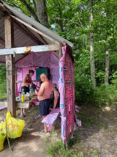 The image depicts an outdoor gathering in a wooded area, possibly a campsite or a rustic shelter. The scene is set during the daytime, with natural light filtering through the trees, suggesting it's likely late morning or afternoon. The weather appears to be warm and possibly humid, given the subjects' attire.
The central focus is on a rough, open-sided wooden structure, possibly a makeshift shelter or a lean-to. The roof appears to be covered with corrugated material, and the walls are draped with pink and white plastic sheeting, one roll featuring repeating text that is difficult to fully decipher but includes stylized logos and what might be product information.
Several individuals are present. A shirtless man, appearing to be in his late 50s or 60s, with a tattoo on his upper arm, is bending over a wooden table. He is wearing dark blue shorts and grey athletic shoes. A woman, likely in her 30s, with her hair pulled back and wearing sunglasses and a red tank top, is standing next to him, also engaged with the table. Her attire suggests casual wear suitable for warm weather. Another person, whose face is partially obscured and wearing a camouflage hat, is seen in the background, also near the table. Their legs are visible, suggesting they are sitting or kneeling.
On the wooden table are various items, including plastic bottles, what appears to be a yellow can or spray bottle, some food items, and possibly some tools or supplies. A wire cage is also visible in the foreground. In the lower right corner, a bright yellow plastic bag with visible text, including Cyrillic characters that read "НАШІ СМАКІ" (Our Tastes), is prominently displayed. This text, along with the location context provided ("Вяззеўскі сельскі Савет, Belarus"), strongly indicates the setting is in Belarus. The overall atmosphere suggests a relaxed, informal gathering, perhaps a picnic or an outdoor work session. The natural setting with trees and vegetation indicates a rural or park-like environment.