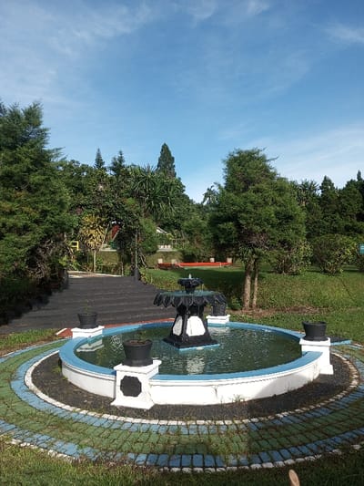 The image depicts an outdoor landscaped area, identified as being in Salai Har Htan, Myanmar. A circular fountain is centrally located in the foreground. It features a multi-tiered, ornamental central structure, predominantly black with white accent panels, from which water cascades into the basin. The fountain basin is white with a light blue inner edge and contains clear water. Four dark-colored planters are visible; two are positioned on white pedestals immediately surrounding the fountain, and two are submerged within the fountain's basin. A paved ring encircles the fountain, exhibiting a pattern of blue, green, and dark grey blocks.

To the left of the fountain, a dark-colored staircase ascends towards an area of dense green foliage. To the right, a grassy slope extends into the midground, populated by scattered evergreen trees and various shrubs. In the background, a dense canopy of mixed trees forms a natural barrier. Beyond this foliage, on a higher elevation, sections of structures with red roofs and white fascias are partially visible. The upper portion of the frame is occupied by a clear, light blue sky with minimal, wispy cloud formations. The scene is illuminated by natural daylight. No individuals or animals are present.