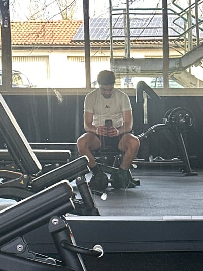 The image depicts the interior of a fitness facility in Jestetten, Germany, with a large window looking out onto an exterior scene. A male subject, appearing to be in his twenties or thirties, is seated on a black padded gym bench, central to the frame. He wears a light-colored t-shirt with a dark logo on the left chest, dark shorts, dark socks, and dark athletic shoes. He is holding a black smartphone with both hands and is looking intently at its screen.
To the subject's right, a black gym machine with a loaded circular weight plate is visible. In the left foreground, components of another black gym machine, including padded sections, are partially visible. The floor is dark grey gym matting.
Through the large window, an exterior environment is visible. A light-colored building with an orange-tiled roof, featuring a prominent array of solar panels, stands in the background. Bare tree branches are discernible against a bright, overcast sky. A metallic structure, possibly a railing or staircase, is also visible outside the window, alongside a partial view of a vehicle. The window surface shows reflections of the gym's interior.
