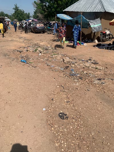 This is a ground-level, outdoor, medium shot of a bustling outdoor market in Dull A, Nigeria. The scene is chaotic and dusty, with a mixture of people, vehicles, and merchandise. A dirt path runs through the middle of the frame, littered with debris and trash.

In the foreground, a person is walking from right to left, wearing a blue and white patterned dress and a headscarf. To their left, another person is walking from right to left, wearing a brightly colored short-sleeved shirt and light-colored pants. Behind them, several other people are visible, some walking and some standing.

Several vehicles are present in the midground, including cars, motorcycles, and what appears to be a truck. Some of the vehicles have luggage strapped to their roofs. Tents and makeshift shelters are set up, providing shade for vendors and shoppers. Merchandise, including clothing and other items, is displayed on the ground and under the shelters.

The background is filled with trees and buildings, suggesting a densely populated area. The sky is clear and blue, indicating a sunny day. The overall impression is one of a lively, but somewhat disorganized, marketplace.