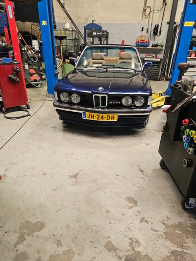 The image depicts the interior of a car repair shop in Bemmel, Netherlands. A dark blue convertible BMW 323i is parked centrally on the concrete floor. The car's headlights are on. In the background, there are two blue hydraulic car lifts, a red tool cart with a drill on it, and various other workshop equipment. A yellow spray bottle hangs from a black tool cabinet on the right side of the image. The overall environment appears organized for vehicle maintenance and repair.
